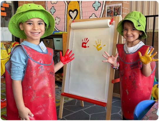 Two preschool girls wearing smocks and green sun hats smiling while finger painting with bright colors at an outdoor easel